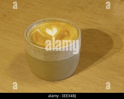 Cappuccino o latte con grafica in schiuma a forma di cuore bianca in tazza in ceramica su una superficie del tavolo in legno Foto Stock