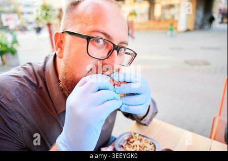 Vista ravvicinata dell'uomo barbuto che morde l'hamburger mentre indossa i guanti blu al caffè all'aperto, osservando la macchina fotografica, il concetto di fast food, l'igiene, il lusso e la moderna cucina urbana Foto Stock