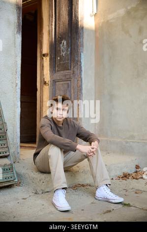 Giovane seduto sulla porta rilassato studente in attesa davanti alla porta infranta indossando un maglione marrone e pantaloni kaki, sneakers bianche, foglie sparse, luce soffusa del tardo pomeriggio, sguardo introspettivo Foto Stock