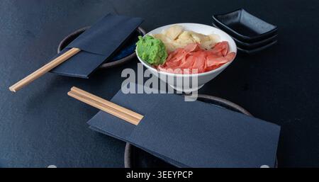 Zenzero bianco e rosa sottaceto e ciotola di wasabi su un tavolo scuro, condimenti tradizionali giapponesi serviti con sushi, piatti con salsa impilata e tovaglioli su BL Foto Stock