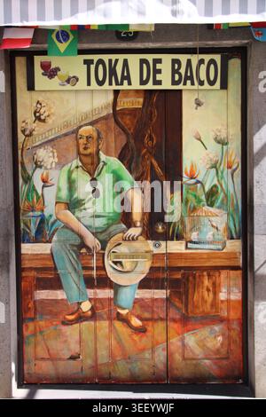 Un vivace murale raffigura un uomo seduto, rilassato e con un cappello in mano. Indossa camicia verde, pantaloni blu, scarpe marroni. Gli occhiali da sole sono appesi al collo. Dietro di lui, piano Foto Stock