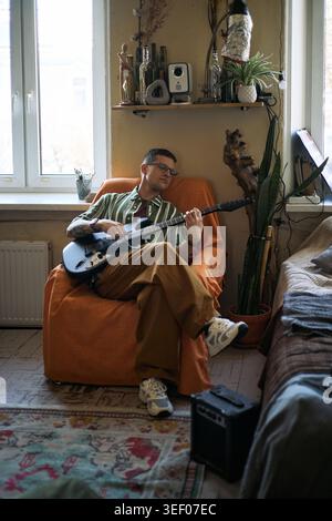 Ripresa verticale di un giovane adulto caucasico che suona la chitarra elettrica mentre sei seduto a casa, mentre pratica musica e ti rilassi. Adatto per hobby, stile di vita creativo e marketing per il tempo libero Foto Stock