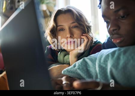 Ritratto di una giovane donna caucasica e di un giovane nero che utilizza un computer portatile e uno smartphone mentre si rilassa a casa, che rappresenta lo stile di vita digitale e la navigazione dei contenuti per il marketing tecnologico Foto Stock