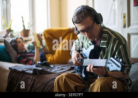 Giovane uomo adulto che indossa le cuffie che suona la chitarra elettrica a casa mentre la donna si rilassa sul letto utilizzando lo smartphone, illustrando l'hobby creativo e il tempo libero a casa per il marketing lifestyle Foto Stock