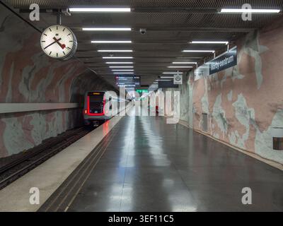 Stoccolma, Svezia - 10 giugno 2025: Moderna piattaforma vuota della stazione della metropolitana con treno fermo, murales, plafoniere e grandi linee analogiche cl Foto Stock