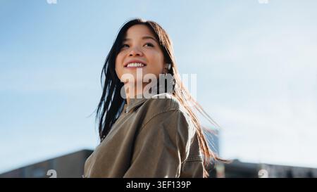 Il ritratto di donna sorridente cattura una giovane donna asiatica che guarda verso il cielo, irradiando fiducia e gioia nella calda luce del sole urbano. Espressione naturale Foto Stock