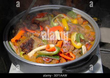 Il vapore sale da una pentola frizzante di verdure vivaci. I peperoni rossi, gialli e verdi aggiungono un colore deciso al piatto. Cipolle saltate e pezzi di carne BL Foto Stock