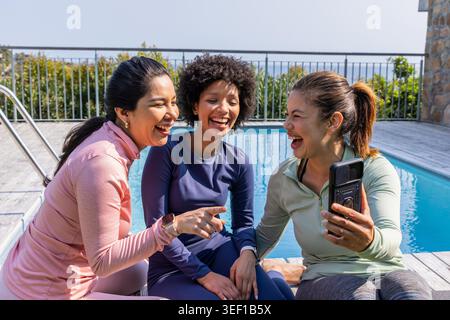 Diverse amiche di abbigliamento sportivo ridono e si appoggiano sul ponte della piscina, tenendo il telefono vicino alla scala. Outdoor, soleggiato, vivace, abbigliamento fitness, athleisure, cameratismo, smartphone Foto Stock