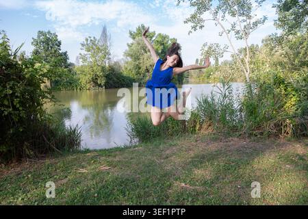 Giovane donna in abito blu si sente felice, saltando con le braccia aperte in un ambiente verde all'aperto Foto Stock