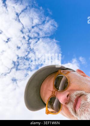 Un ritratto verticale ad angolo basso di un anziano con una barba grigia che indossa occhiali da sole e un berretto contro un cielo blu con nuvole bianche Foto Stock