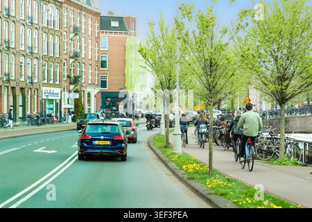 Amsterdam, Paesi Bassi - 17 aprile 2026: Vivace scena urbana con auto, motociclette e ciclisti su una pista ciclabile dedicata lungo un canale, fiancheggiato da wi Foto Stock
