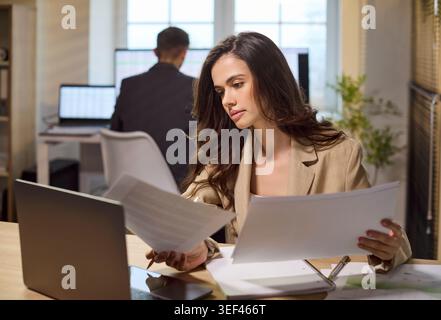 Donna d'affari concentrata che esamina documenti e lavora su un notebook in un ufficio moderno Foto Stock