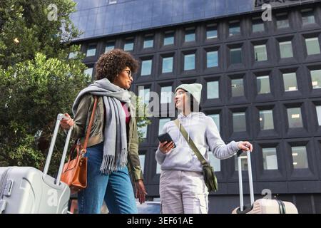 Due donne che camminano e parlano sul marciapiede della città con valigie rotanti e smartphone Foto Stock