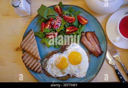 Autentica composizione piatta di un sostanzioso pasto mattutino. Un rustico piatto blu comprende uova fritte con il lato soleggiato, strisce di pancetta, toast alla griglia e un Foto Stock