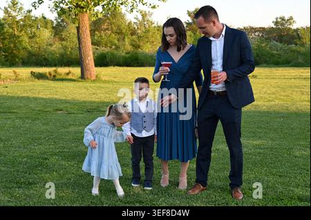 Coppia matura con due bambini in piedi sull'erba durante la festa in giardino e che tengono cocktail Foto Stock