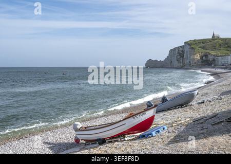 Vista della spiaggia di ciottoli di Etretat con una piccola barca di legno rossa e bianca e un gommone gonfiabile in primo piano al largo delle caratteristiche scogliere del Foto Stock