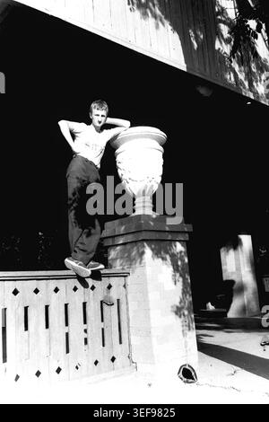 Sloviansk, 1983 anni. Un giovane uomo colpisce una posa sicura sul terreno della stazione termale di Slavkurort. In piedi su una recinzione decorativa in pietra, si appoggia casualmente contro un grande vaso bianco classico, con una mano appoggiata dietro la testa. Indossa una t-shirt leggera, pantaloni scuri e scarpe da ginnastica. L'illuminazione ad alto contrasto crea ombre spettacolari sotto la sporgenza in legno dell'edificio. Questo elegante ritratto documentario conserva splendidamente lo stile di vita spensierato delle vacanze e lo spirito giovanile del tranquillo Donbas. Foto Stock