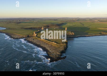 Vista aerea delle rovine del castello di Dunstanburgh su un promontorio roccioso che si affaccia sul Mare del Nord con campi verdi e onde che si infrangono Craster, Inghilterra, U Foto Stock