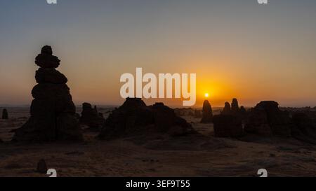 Il cairn di pietra e il pinnacolo di arenaria si stagliano contro il vivace tramonto arancione sulla pianura desertica, nella regione di Alula, Arabia Saudita Foto Stock