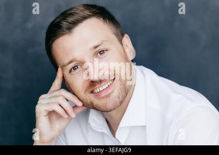 Un dipendente maschio allegro ha un'espressione gioiosa, tiene il dito sul tempio, si rallegra per iniziare la vacanza, vestito con una camicia bianca formale, posa su sfondo scuro. Uomo d'affari di successo al coperto. Foto Stock