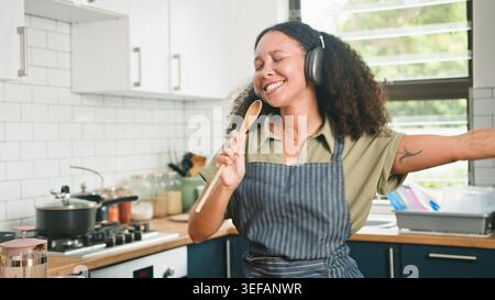 Donna allegra che canta e balla in cucina mentre cucina con le cuffie e il cucchiaio di legno Foto Stock