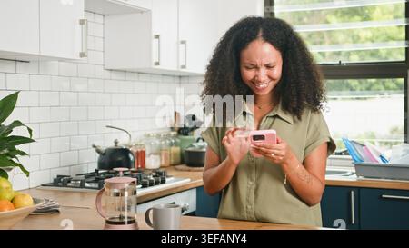 Donna felice che usa lo smartphone in Bright Kitchen mentre prepara il caffè e sorride al messaggio Foto Stock