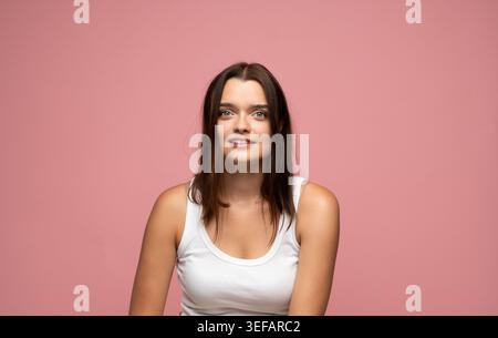 Giovane donna bruna che guarda la fotocamera con un'espressione amichevole, indossa una canotta bianca su sfondo rosa Foto Stock
