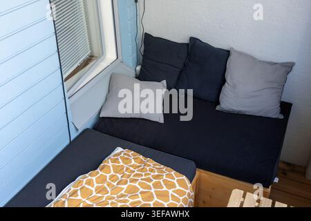 Una panca in legno del balcone e' dotata di cuscini scuri, vari cuscini grigi e una coperta a fantasia giallo brillante per creare una comodita' Foto Stock