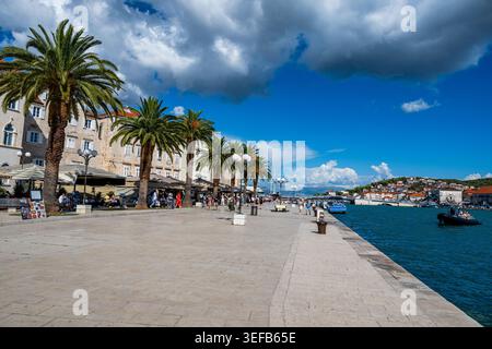 I visitatori passeggiano lungo il lungomare di Traù mentre le barche passano sullo sfondo in una giornata di sole Foto Stock