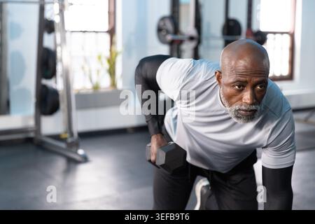Uomo afroamericano anziano che si esibisce in una fila pendente in palestra, tenendo un manichino esagonale, indossando abbigliamento sportivo. Fitness, pesi, allenamento, forza, luce del sole, stu Foto Stock