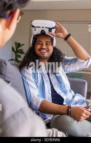 Amici seduti su un divano grigio in salotto, sollevando il visore VR bianco, sorridendo, indossando un orologio da polso. Gadget, lounge, casa, moderno, tecnologia, casual, intera Foto Stock