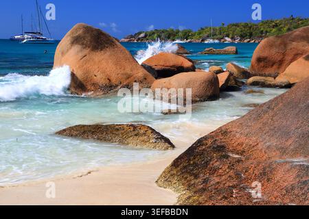 Le onde si schiantano contro la costa rocciosa di Anse Georgette sull'isola di Praslin con le barche sullo sfondo Foto Stock