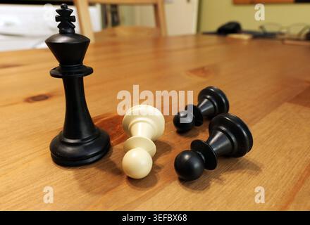 Il pezzo nero del re degli scacchi si erge alto su un tavolo di legno, guardando attraverso un pedone bianco caduto e due pedine nere cadute, simboleggiando la vittoria, la sconfitta, A. Foto Stock
