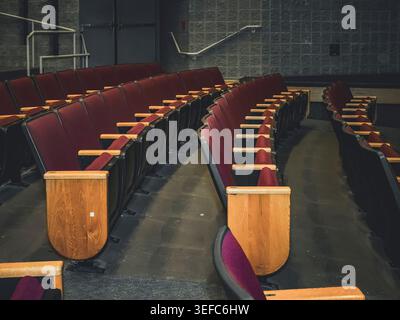 File di posti a sedere vuoti rivestiti di rosso con pannelli di legno in un auditorium vuoto con pareti in mattoni di cemento e porte di uscita Foto Stock