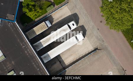 Vista aerea dei semi-camion bianchi parcheggiati presso le banchine di carico di un magazzino industriale con alberi verdi e strade asfaltate a Nieuwegein, Utrecht, Netherlan Foto Stock
