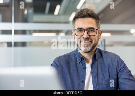 Uomo imprenditoriale con barba e occhiali che guardano la fotocamera dalla scrivania, rappresentando fiducia, successo e una visione positiva in un ambiente aziendale contemporaneo Foto Stock