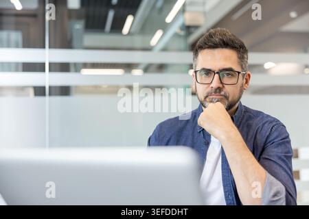 Uomo medio adulto con barba e occhiali che guardano intensamente la macchina fotografica, mano sul mento, brainstorming idee durante una giornata di lavoro in un moderno ambiente d'ufficio Foto Stock