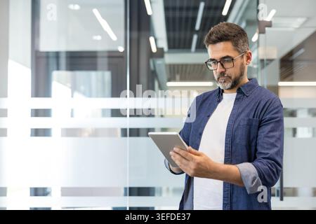 Uomo d'affari con il barbuto che indossa occhiali e una camicia in denim che si impegna attivamente con un tablet digitale, gestisce gli affari e si connette in un ambiente d'ufficio contemporaneo Foto Stock