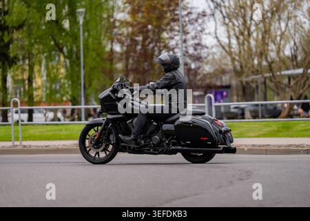 Vista laterale di un motociclista in pelle nera che guida una potente motocicletta da turismo nera su una strada urbana con alberi verdi di parco sullo sfondo represen Foto Stock