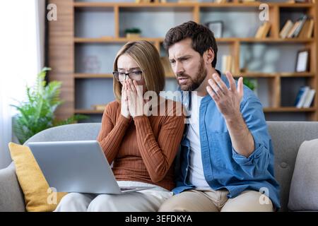 Una coppia preoccupata, seduta su un divano, soffre di stress e shock mentre guarda lo schermo di un notebook, leggendo cattive notizie sulle difficoltà finanziarie e sul debito Foto Stock