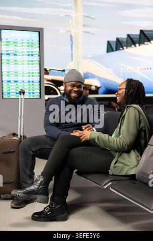 Amanti afroamericani seduti nella sala del terminal dell'aeroporto con vista sulla pista, tenendosi per mano mentre discutono dell'itinerario e dei programmi di vacanza. Coppia che viaggia insieme, godendo del tempo libero. Foto Stock