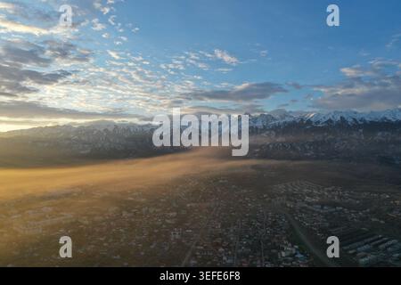 Una prospettiva aerea cattura un'estesa cittadina di montagna annidata sotto torreggianti vette innevate mentre la calda luce del sole dorata illumina la valle flo Foto Stock