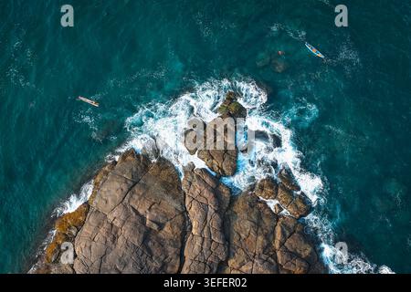 Vista aerea del promontorio roccioso con onde che si infrangono contro la riva e piccole barche da pesca nel mare turchese di Kovalam, Kerala, India. Foto Stock