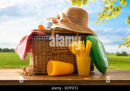 Cestino da picnic in vimini con cappello, occhiali da sole e stoviglie colorate su un soleggiato tavolo di campagna sotto il cielo blu e foglie verdi. Foto Stock