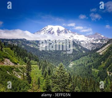 Il Monte Rainier, coperto di neve, si riflette in un tranquillo lago alpino circondato da una foresta sempreverde sotto un cielo azzurro Foto Stock