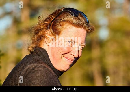 Una donna di mezza età con i capelli rossi che si insinuano nel sole mentre guarda la fotocamera Foto Stock