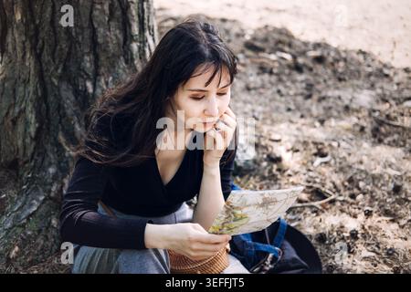 La persona si siede sotto l'albero mentre legge attentamente la mappa cartacea nella foresta. Leggere la cartina cartacea, trovare la strada, Lost in the Woods Concept, pianificare il percorso Foto Stock