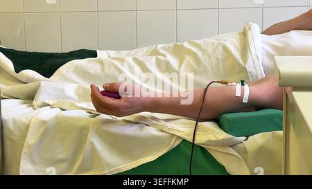 Persona sdraiata sul letto medico sottoposta a plasmaferesi, donando plasma ematico via iv nel braccio mentre stringe una palla da stress, personale medico e tubo di vista Foto Stock