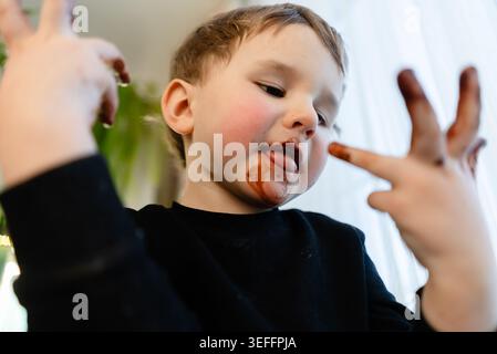 Un bambino di tre anni sta per leccare il cioccolato fuso dalle dita. Concentrati su mani e bocca. Concetto di dessert proibito, abitudini alimentari malsane, allergie e diabete. Foto Stock