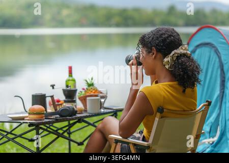 Donna nera che scatta foto con la macchina fotografica mentre campeggia in riva al lago in mezzo alla natura Foto Stock